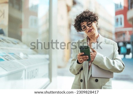 Woman with laptop and smartphone, standing on street of city centre, smiling at camera. Business woman leader wearing suit standing in big city using cell phone platform applications.