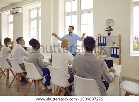 Young cheerful and confident man makes presentation about business ideas in front of business committee. Man speaks and gestures in front of people sitting in row in bright big boardroom. Royalty-Free Stock Photo #2309837271