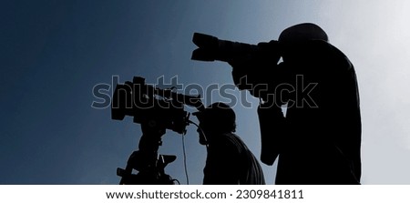 Camera operator on a tripod, photographer, cameraman silhouette isolated on blue sky background. 2 two men working together. 