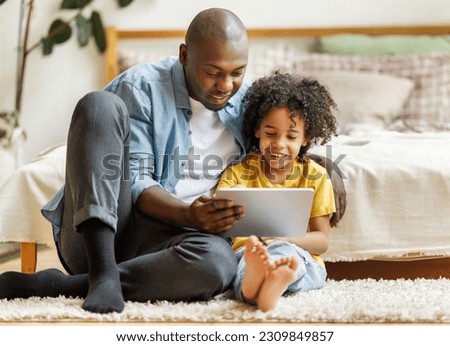 Happy african american family little boy son with young dad watching funny videos on tablet together at home, father with child using computer in bedroom while enjoying weekend
