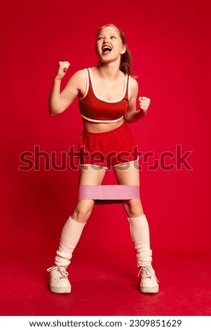 Gluteal muscles workout. Happy sporty girl wearing sportswear and doing exercise with rubber bands for sport over red background. Concept of sport, hobby, fitness, fashion, emotions, beauty, ad Royalty-Free Stock Photo #2309851629