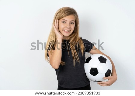 Happy young caucasina girl holding a football ball over white background touches both cheeks gently, has tender smile, shows white teeth, gazes positively straightly at camera,