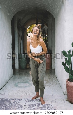Full body of happy young female standing in narrow entrance passage with green potted plants and looking at camera while holding stomach with hand in sunlight and enjoying summer vacation Royalty-Free Stock Photo #2309852427