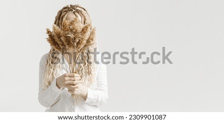 Woman holding pampas grass in front of face, light background, autumn, fall minimal trend concept. Female with long hair dreadlocks hiding behind bouquet dried plant. Creative portrait photo ideas