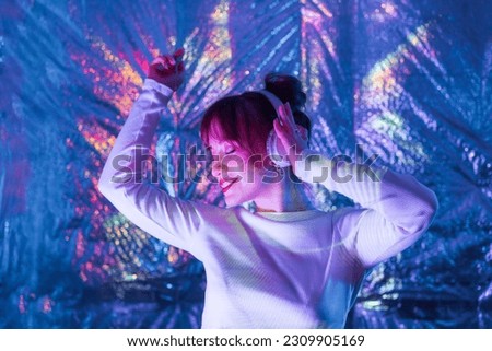 Relaxed smiling woman with closed eyes in white clothes and headphones dancing in neon light. Music lover. White dress code party. Silent disco. Enjoy moment on shiny background. Selective focus. Royalty-Free Stock Photo #2309905169