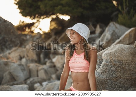 Cute young girl sits on the sea rock, posing and grabs the last rays of the sunset sun.