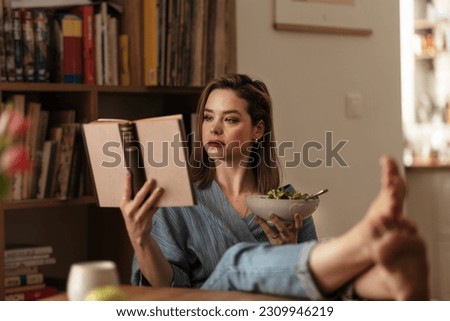 Woman resting at home, reading book and eating salad. Royalty-Free Stock Photo #2309946219
