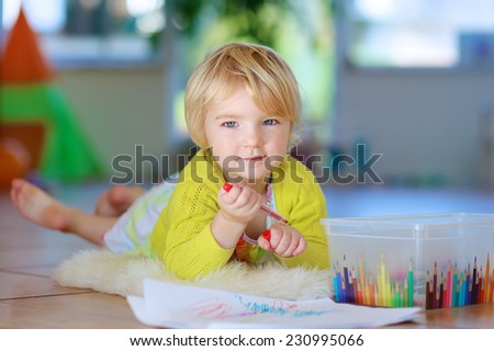 Happy little child, adorable blonde toddler girl lying comfortable on tiles floor on warm lambskin drawing on paper with colorful pens on sunny day