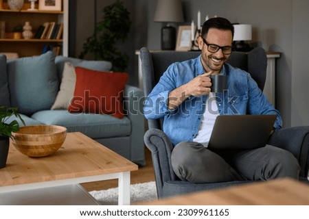 Smiling young businessman drinking coffee and reading e-mails over wireless computer while sitting on armchair. Happy male freelancer having drink and working remotely over laptop at home office Royalty-Free Stock Photo #2309961165