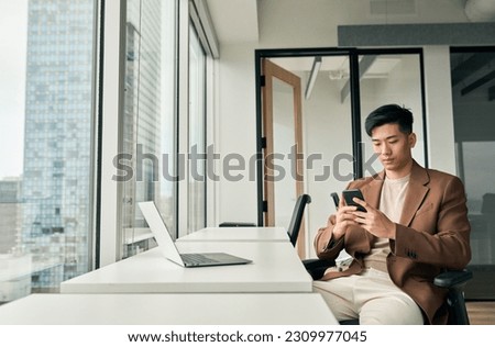 Young busy Asian business man manager using laptop and mobile phone tech in office. Professional Japanese businessman holding smartphone, working on cellphone looking at cell sitting at desk.