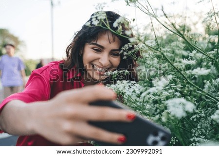 Selective focus of delightful ethnic woman taking self portrait on smartphone against green bush with small white flowers in summer