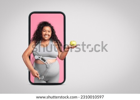 Smiling black plus size woman in sportswear with scales and apple on smartphone screen isolated on grey studio background, banner, free space. Body care, sports, app for weight loss and fitness