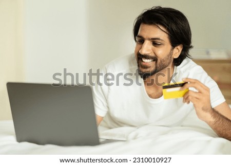Positive millennial middle eastern guy lying on bed with credit card and laptop, enjoy good morning, online shopping in bedroom interior. Order remotely, cashback, sale, ad and offer