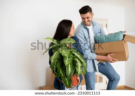 Moving Concept. Portrait Of Cheerful Young Couple With Belongings Posing In Their New Home, Happy European Spouses Holding Cardboard Box And Plant And Smiling, Relocating To New Home, Copy Space Royalty-Free Stock Photo #2310011113