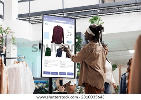 Customer using touch display shopping for fashion items in clothing store, interactive board. African american man choosing stylish clothes with different designs, modern kiosk service. Royalty-Free Stock Photo #2310035643
