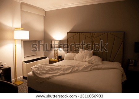 Dark interior of an luxury hotel room at night with lamps and an unmade bed with four pillows and a queen sized bed.  Funny video conference background or for travel advertisements. Royalty-Free Stock Photo #2310056903