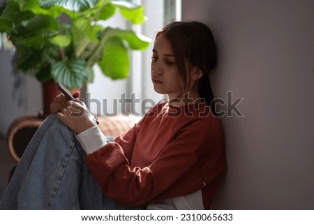 Sad depressed teen adolescent girl looking side hold phone sit on floor at home. Upset teenager with cellphone waiting for call feel bad anxious need help. Cyberbullying, difficult relations at school Royalty-Free Stock Photo #2310065633