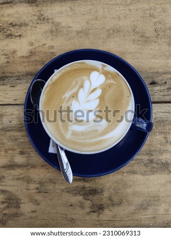 A cup of seductively scented coffee adorns the wooden table with radiating warmth. The pretty blue cup and saucer add visual interest, while the latte art on the coffee surface embodies the barista's 