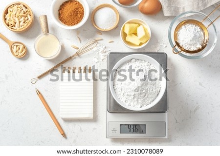 Flour in bowl on digital scale with notepad and pen and baking ingredients, flour, nuts, eggs, sugar, milk and gold whisk on marble table, flatlay Royalty-Free Stock Photo #2310078089