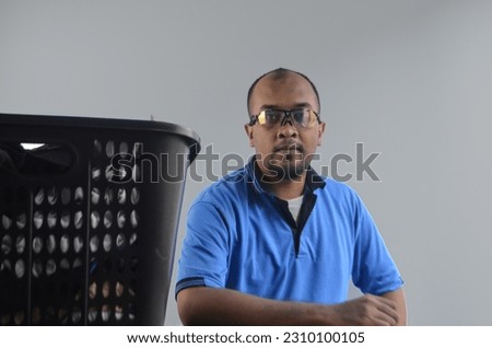 asian man wearing blue shirt and glasses sitting near basket of clothes