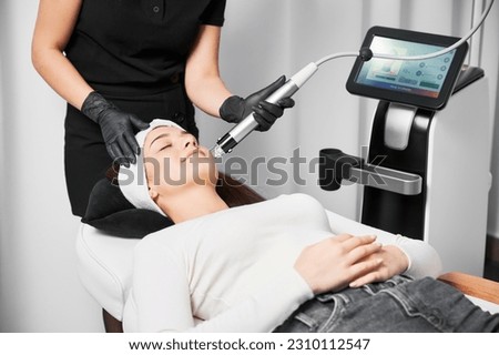 Doctor beautician using radiofrequency microneedling device while performing face lifting procedure for patient. Woman lying near cosmetology equipment and receiving skincare rejuvenation treatment. Royalty-Free Stock Photo #2310112547