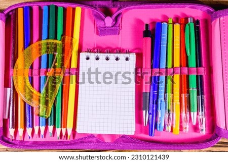 Different school stationeries (pens, pencils, notepad and protractor) in pink pencil box. Top view