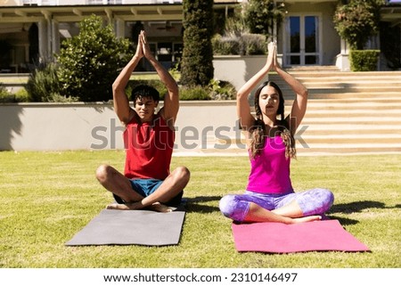 Biracial young couple with arms raised practicing meditation while sitting on mats in yard. Unaltered, love, togetherness, summer, zen, yoga, fitness and active lifestyle concept.
