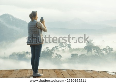 A woman photographs on a smartphone a beautiful mountain range in foggy clouds in the early summer morning.