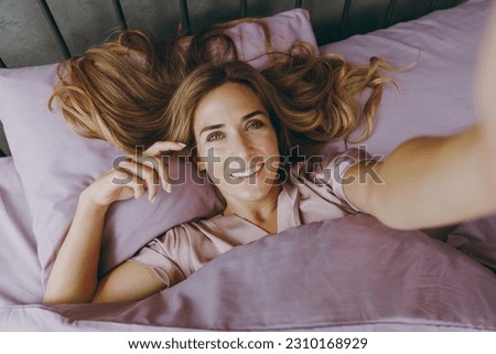 Young woman wear purple t-shirt pajama lying in bed doing selfie shot pov on mobile cell phone rest relax spend time in bedroom lounge home in own room hotel wake up dream be i good mood day. concept