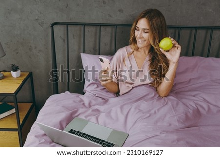 Young IT woman wear t-shirt pajama sits on bed hold use work on laptop pc computer use mobile phone eat apple rest relax spend time in bedroom lounge home in own room hotel wake up be in good mood day