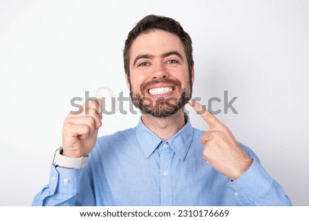 Businessman wearing blue t-shirt over white background holding an invisible aligner and pointing perfect straight teeth. Dental healthcare and confidence concept.