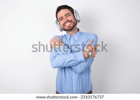 Charming pleased business man wearing blue t-shirt with headphones over white background embraces own body, pleasantly feels comfortable poses. Tenderness and self esteem concept