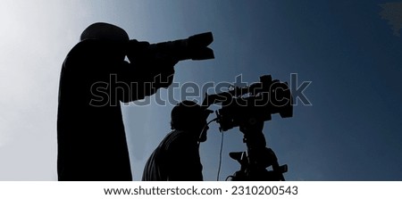 Camera operator on a tripod, photographer, cameraman silhouette isolated on blue sky background. 2 two men working together.  Royalty-Free Stock Photo #2310200543