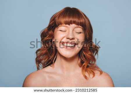 Happy woman with ginger hair and flawless skin smiling with her eyes closed. Cheerful young woman embracing her natural beauty. Body confident young woman standing against a blue studio background.