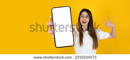 Pointing finger mobile phone, portrait of young caucasian excited girl pointing finger mobile phone. Blank empty white big screen smartphone mockup. Isolated yellow studio background, copy space.  Royalty-Free Stock Photo #2310234573