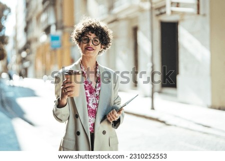 Dreamy woman in formal clothes carries notepad tablet disposable cup of coffee has walk outdoor during daytime poses near office building with cheerful expression going to have job interview