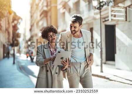 Business people outdoors. Handsome business man and his beautiful female colleague discussing new project while crossing the street, urban background.  Two colleagues using modern gadgets for work 