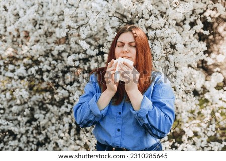 Sneezing young redhead woman with nose wiper among blooming trees in park. Portrait of sick women sneezes in white tissue, suffers from rhinitis and running nose. Symptoms of cold or allergy. Royalty-Free Stock Photo #2310285645