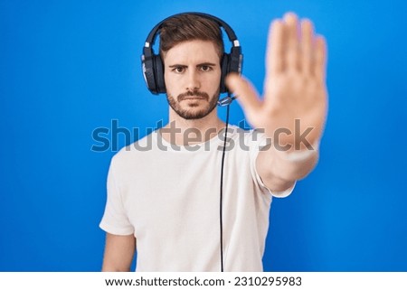 Hispanic man with beard listening to music wearing headphones doing stop sing with palm of the hand. warning expression with negative and serious gesture on the face. 