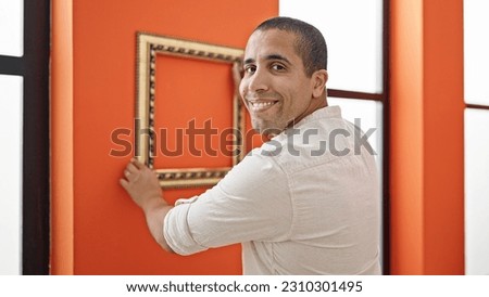 Young hispanic man smiling confident hanging retro empty frame on wall at home
