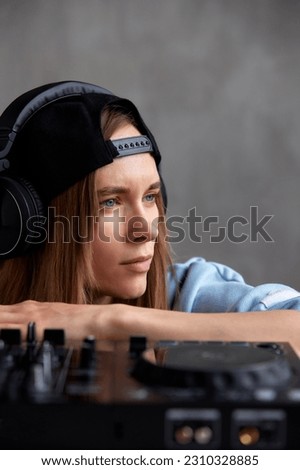 A young pretty long haired DJ girl in a blue sweater and a black baseball cap poses with a black DJ mixing console. Close-up studio shot, gray background.