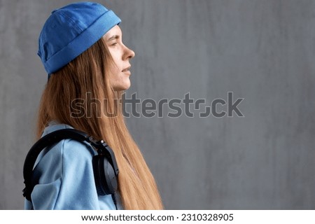 A young pretty long-haired girl in a blue sweater and a funny hat with black DJ headphones on her shoulder. Music, fun and relaxation. Close-up studio shot, gray background.