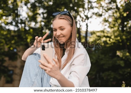 Pensive blonde woman make selfie posing on park background. Outdoor shot of happy business lady show peace sign. Business freedom style. Girl raises her hand to camera, hold mobile, v gesture.