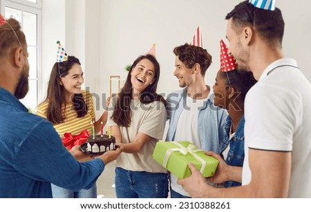 Multiracial group of excited friends in festive hats giving a cake and present gift to a happy young woman at her birthday party at home. Young brunette woman in her 20s getting presents having fun.