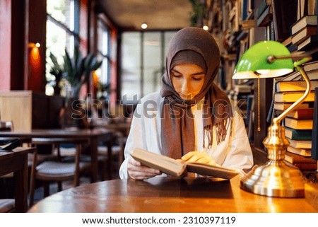 Serious Islamic teen girl in hijab reading book at library. Thoughtful muslim female teenager adolescent in headscarf and white shirt looking through textbook and preparing for the exam indoors.