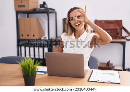 Young blonde woman working at the office wearing glasses smiling happy doing ok sign with hand on eye looking through fingers 
