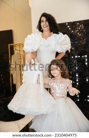 charming middle eastern bride with brunette hair standing in white wedding gown with puff sleeves and ruffles and holding girly dress with tulle skirt near daughter in bridal store