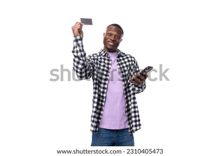positive smiling young african man with short haircut in plaid shirt holding card mockup and phone on white background with copy space