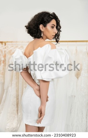 stunning middle eastern bride with brunette and wavy hair posing in stylish wedding dress with puff sleeves and ruffles in bridal boutique next to tulle fabrics, elegant woman