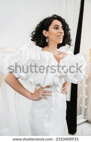 positive middle eastern woman with brunette and wavy hair posing with hands on hips in trendy wedding dress with puff sleeves and ruffles in bridal salon next to tulle fabrics